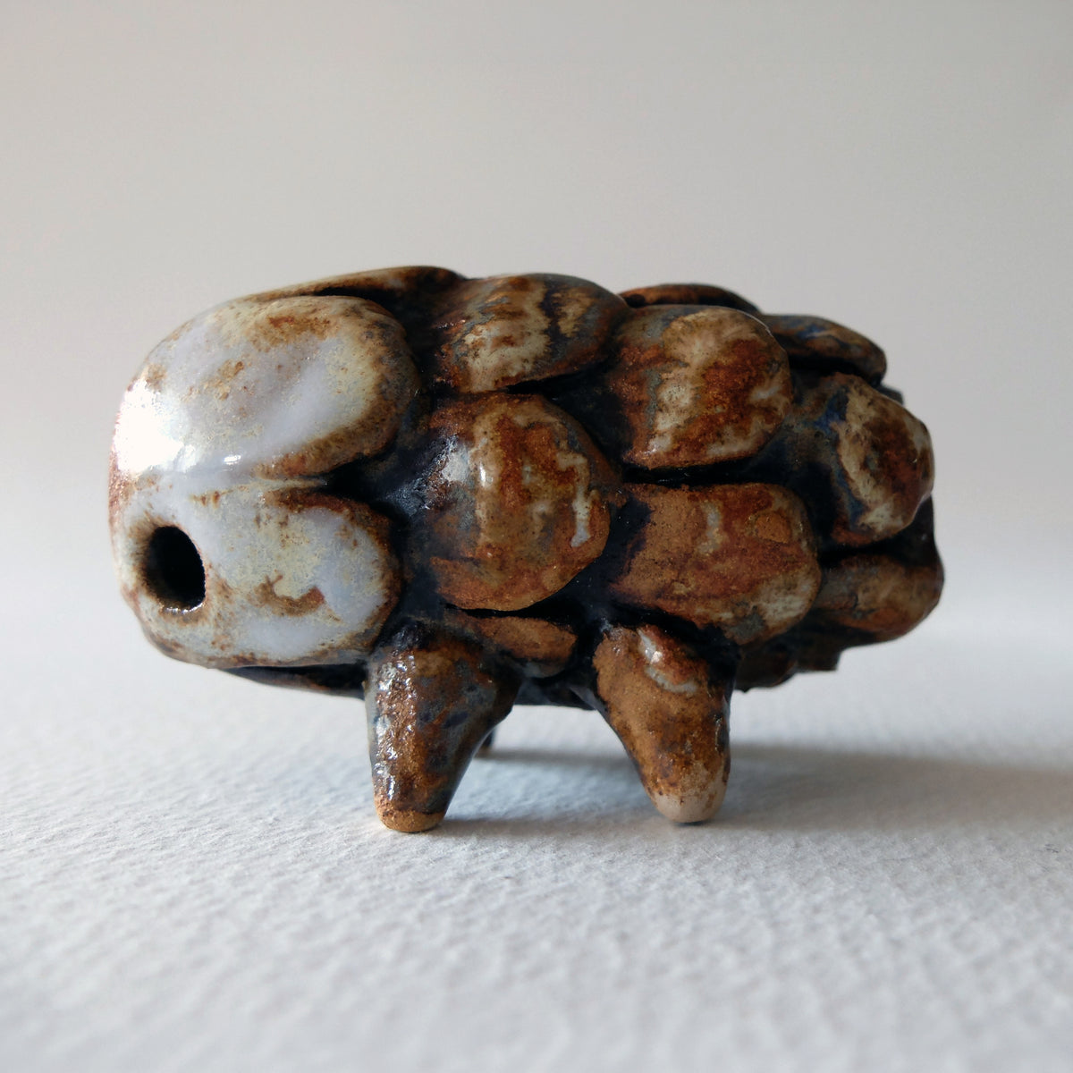 Handcrafted ceramic figurine titled The Pinecone Hedgehog »Igel-Kotten« with a pinecone scale texture, standing on four small legs in a glossy brown, cream, and dark blue-grey glaze, sculpted by Nikolaj Jesper Cyon.