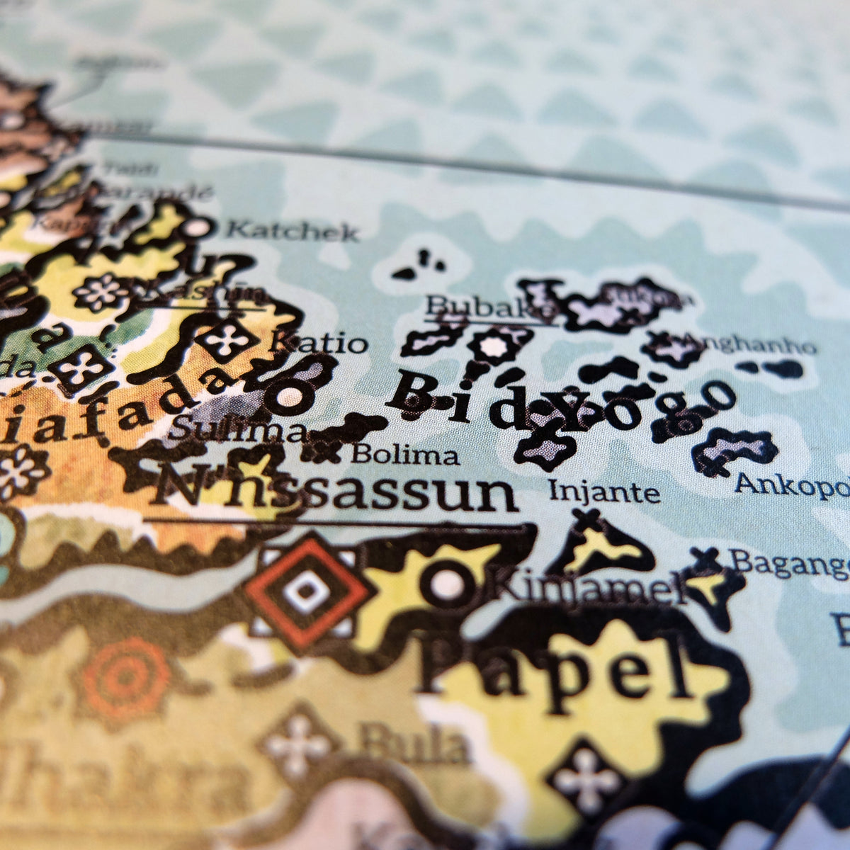 Macro photograph showing the Bidyogo archipelago (Bijagós Islands) and the mainland coastline on the Alkebu-Lan map print. Features island kingdoms including Bubake and Injante alongside coastal realms such as Biafada and Papel, with its royal capital N'nssassun (today known as Bissau), printed on textured paper by Nikolaj Jesper Cyon.