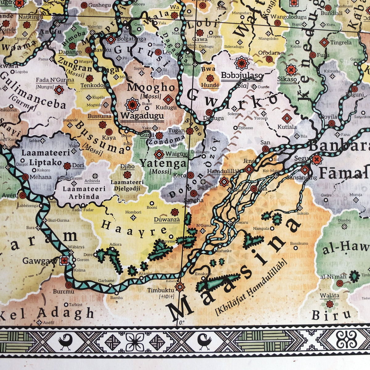 Macro photograph showing the Niger River bend and Sahel region on the Alkebu-Lan map print, covering modern Mali and Burkina Faso. Features Maasina, Gawgaw [Gao], Mossi realms like Wagadugu, Tuareg territories, and the Inland Niger Delta. Here, the prime meridian (0°) runs through Timbuktu and the Sankore University instead of Greenwich/Oxford in our world. Below, the decorative border includes an Adinkra Sankofa bird symbol dropping its egg, printed on textured paper by Nikolaj Jesper Cyon.