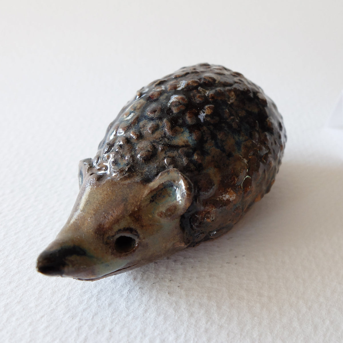 Handcrafted ceramic hedgehog figurine "The Cautious Mulberry Hedgehog (#17)" in stoneware by Nikolaj Jesper Cyon. Features a timid posture with a bumpy, dark brown and charcoal mulberry-textured back and a smooth beige snout.
