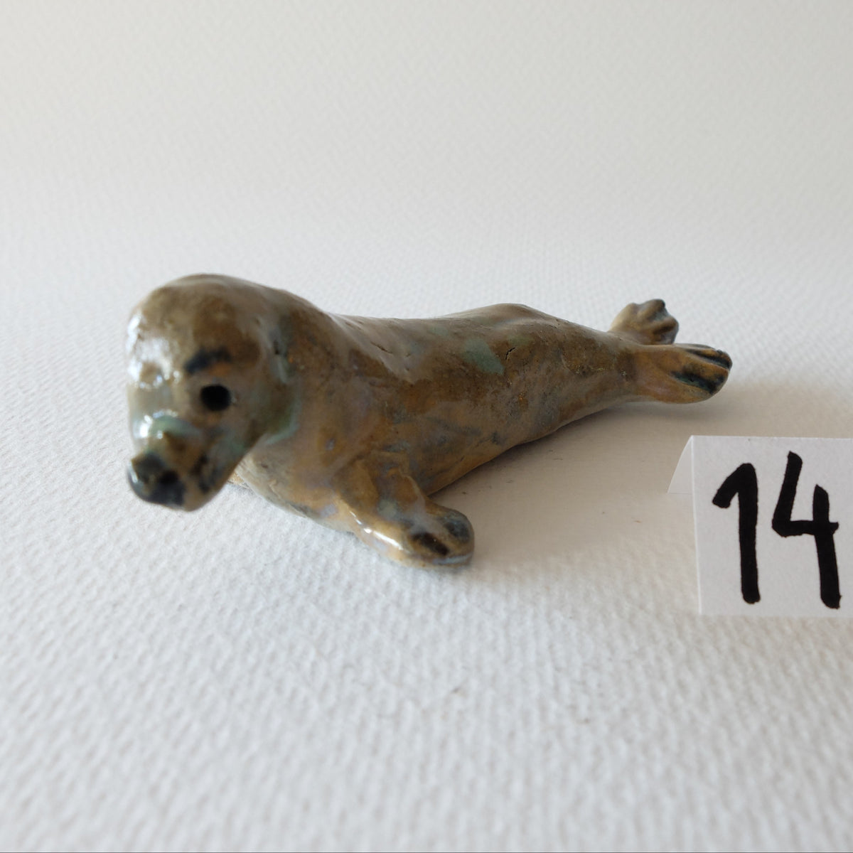 Handcrafted ceramic seal figurine "The Contemplative Seal (#14)" in stoneware by Nikolaj Jesper Cyon. Depicts a seal resting on its belly with raised head and hind flippers, glazed in soft earthy brown, beige, and subtle green tones with dark facial details.