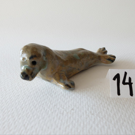 Handcrafted ceramic seal figurine "The Contemplative Seal (#14)" in stoneware by Nikolaj Jesper Cyon. Depicts a seal resting on its belly with raised head and hind flippers, glazed in soft earthy brown, beige, and subtle green tones with dark facial details.