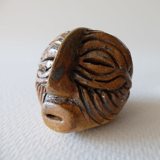 Handcrafted ceramic mask sculpture "The Kifwebe Head (#11)" in stoneware by Nikolaj Jesper Cyon. Features a stylized face inspired by traditional Songye Kifwebe masks, with deep incised parallel grooves, a prominent central nose ridge, elongated eye slits, and a warm earthy brown glaze finish.