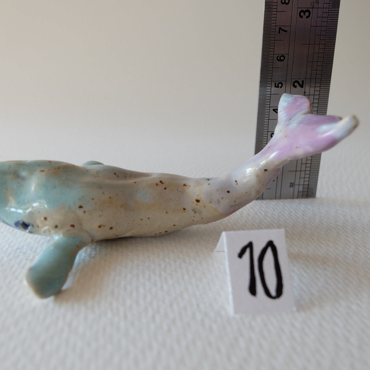 Handcrafted ceramic whale figurine "The Pastel Whale (#10)" in stoneware by Nikolaj Jesper Cyon. Side view with a vertical ruler measuring the height of the raised tail fin.
