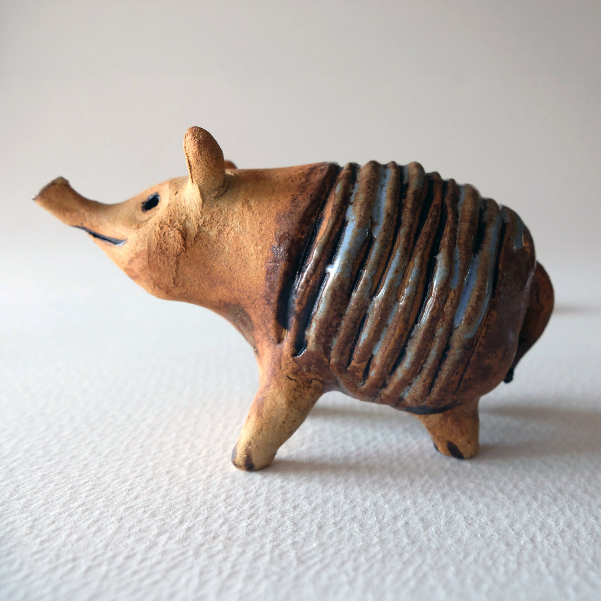 Handcrafted ceramic armadillo figurine with a long snout, upright ears, and a ribbed segmented shell in a contrasting matte terra-cotta and glossy blue-brown glaze, sculpted by Nikolaj Jesper Cyon.