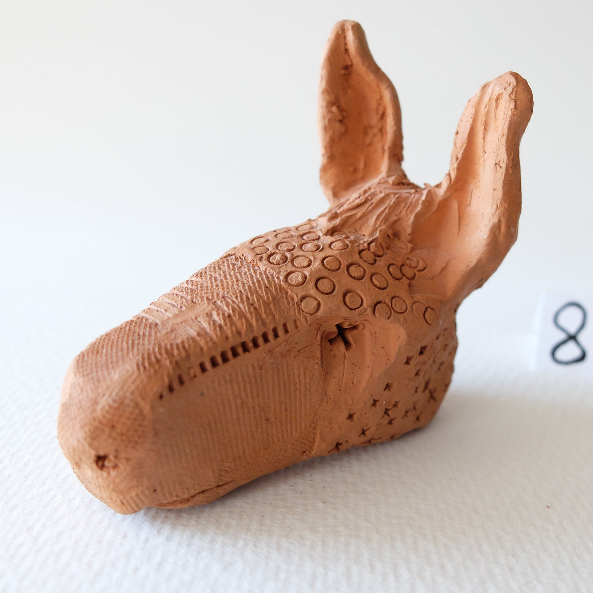 Handcrafted terracotta sculpture "The Babylonian Donkey (#8)" by Nikolaj Jesper Cyon. The unglazed reddish-brown clay sculpture features a donkey's head with long upright ears, detailed with intricate decorative patterns such as small stamped circles, dotted textures, and incised geometric lines.