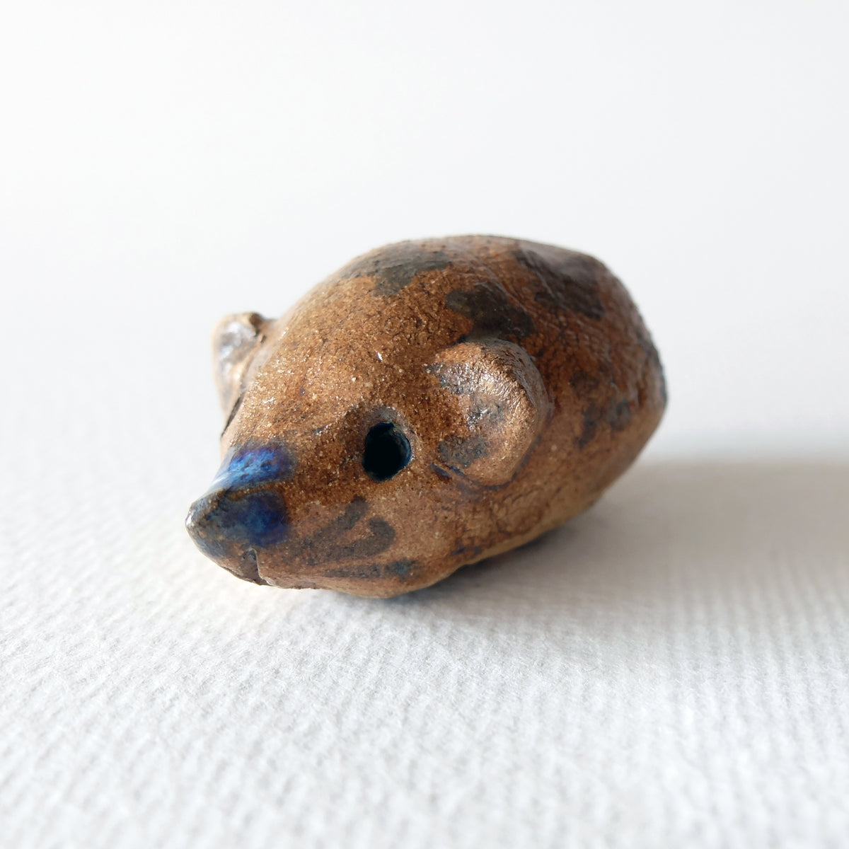 Handcrafted ceramic hedgehog figurine "The Chestnut Hedgehog (#5)" in stoneware by Nikolaj Jesper Cyon. Features a warm, speckled brown and ochre body with dark decorative spots and a touch of blue glaze on the pointed snout, captured in a three-quarter front view.