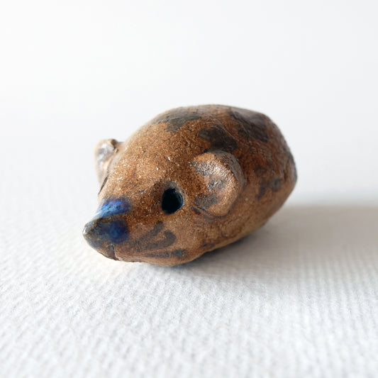 Handcrafted ceramic hedgehog figurine "The Chestnut Hedgehog (#5)" in stoneware by Nikolaj Jesper Cyon. Features a warm, speckled brown and ochre body with dark decorative spots and a touch of blue glaze on the pointed snout, captured in a three-quarter front view.