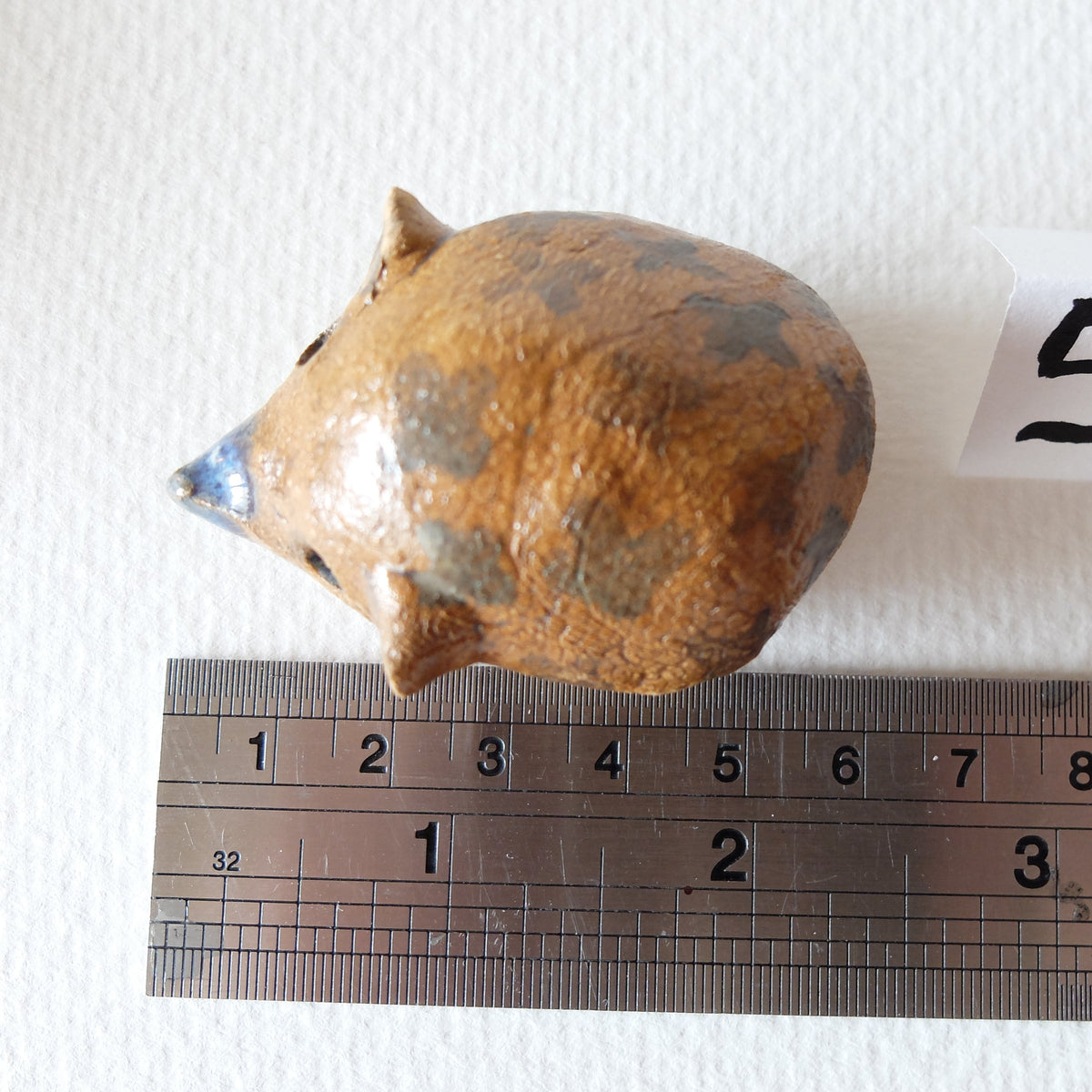 Handcrafted ceramic hedgehog figurine "The Chestnut Hedgehog (#5)" in stoneware by Nikolaj Jesper Cyon. Top-down view next to a metal ruler showing size scale.