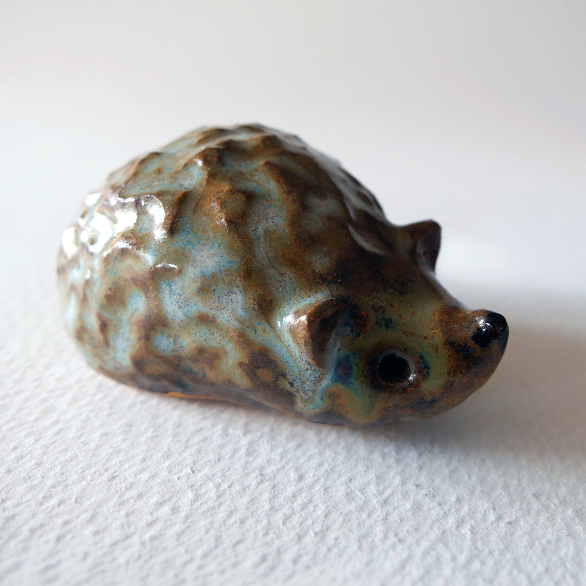 Handcrafted ceramic hedgehog figurine with a knobby textured shell in a mottled pale green-blue and brown glaze, sculpted by Nikolaj Jesper Cyon.