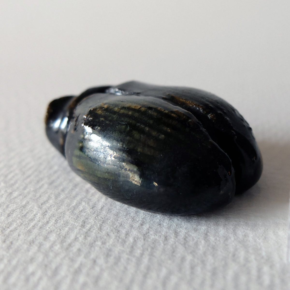 Handcrafted ceramic beetle figurine "The Obsidian Beetle (#4)" in stoneware by Nikolaj Jesper Cyon. Side perspective view showing its dark, glossy carapace glaze and head structure.