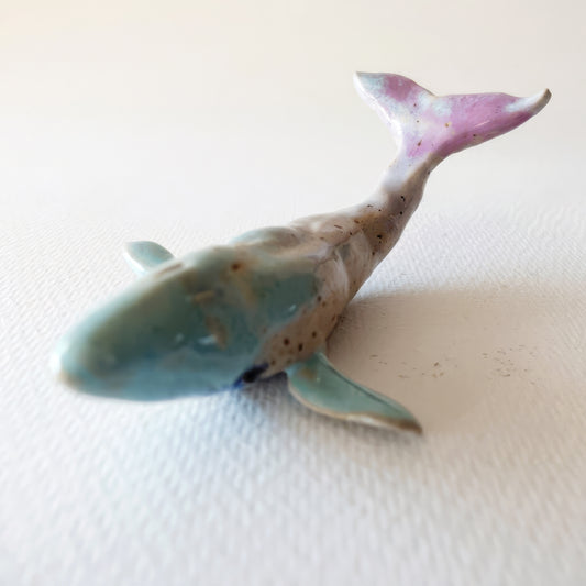 Handcrafted ceramic whale figurine "The Pastel Whale (#10)" in stoneware by Nikolaj Jesper Cyon. Glazed in soft pastel tones with a light blue-green body, speckled beige back, and a raised lavender-pink tail fin.