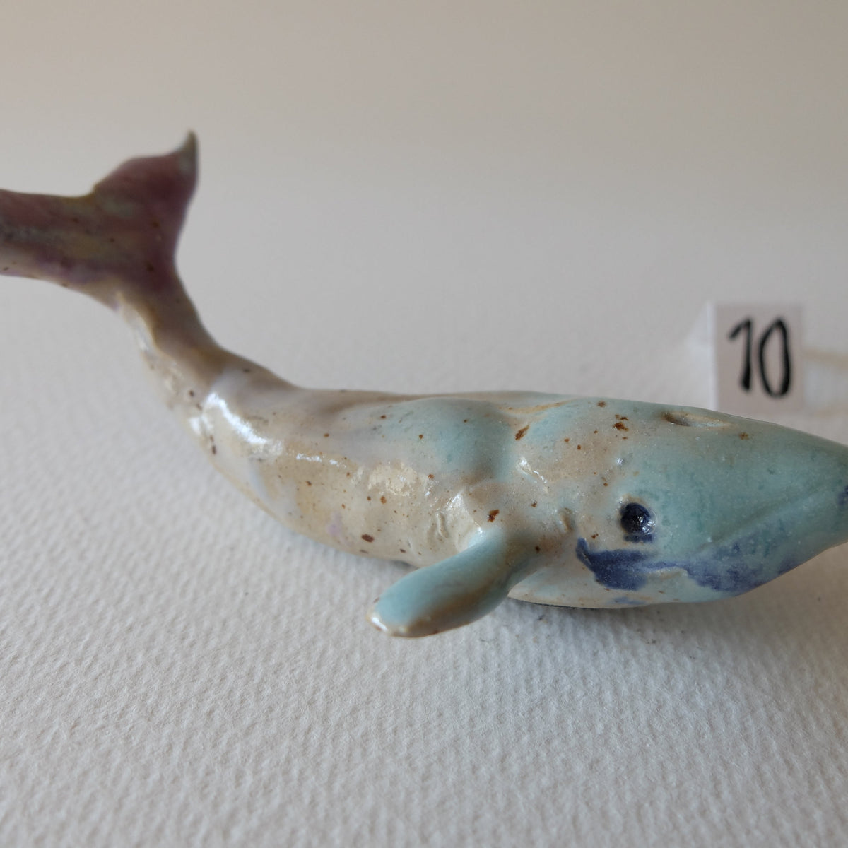 Handcrafted ceramic whale figurine "The Pastel Whale (#10)" in stoneware by Nikolaj Jesper Cyon. Side profile view showing the soft color transition from light turquoise body to a pastel pink tail fin.