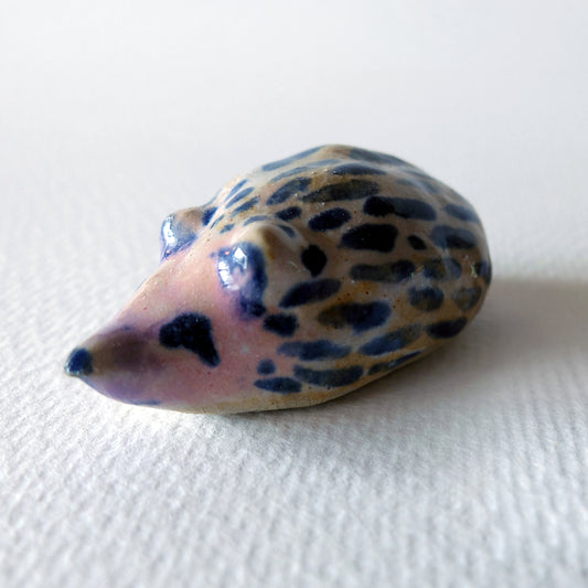 Handcrafted ceramic hedgehog figurine "The Striped Hedgehog (#3)" in stoneware by Nikolaj Jesper Cyon. Small hedgehog sculpture with dark blue stripes and speckled patterned glaze on a light tan and subtle pinkish base.