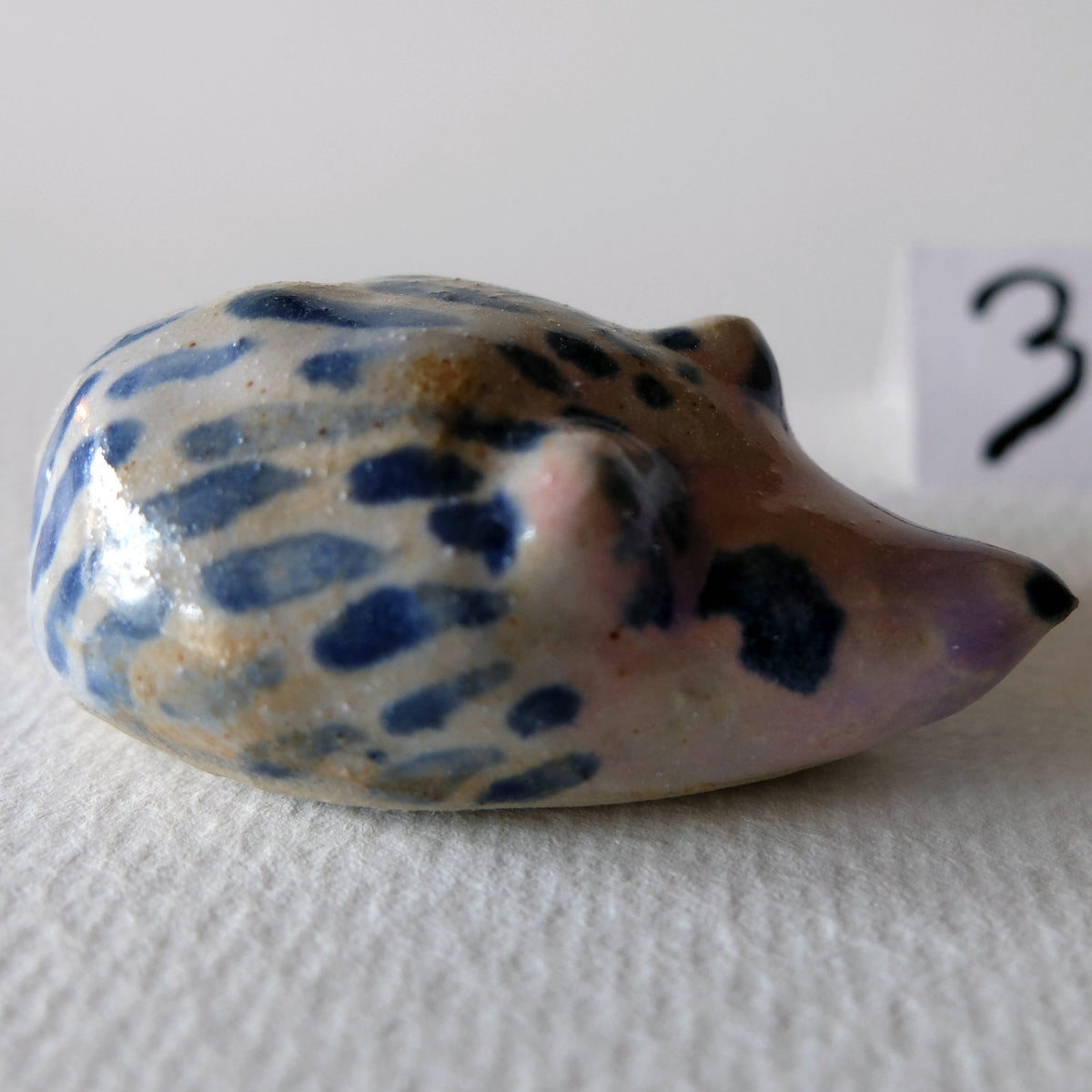 Handcrafted ceramic hedgehog figurine "The Striped Hedgehog (#3)" in stoneware by Nikolaj Jesper Cyon. Side profile view showing its snout, ears, and blue-striped pattern, with a small "3" label in the background.