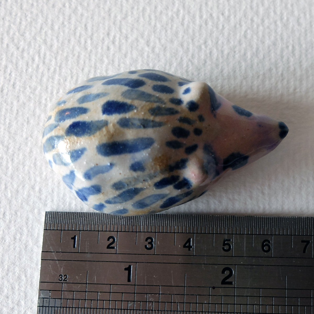 Handcrafted ceramic hedgehog figurine "The Striped Hedgehog (#3)" in stoneware by Nikolaj Jesper Cyon. Top-down view next to a metal ruler to show the size scale of the small sculpture.