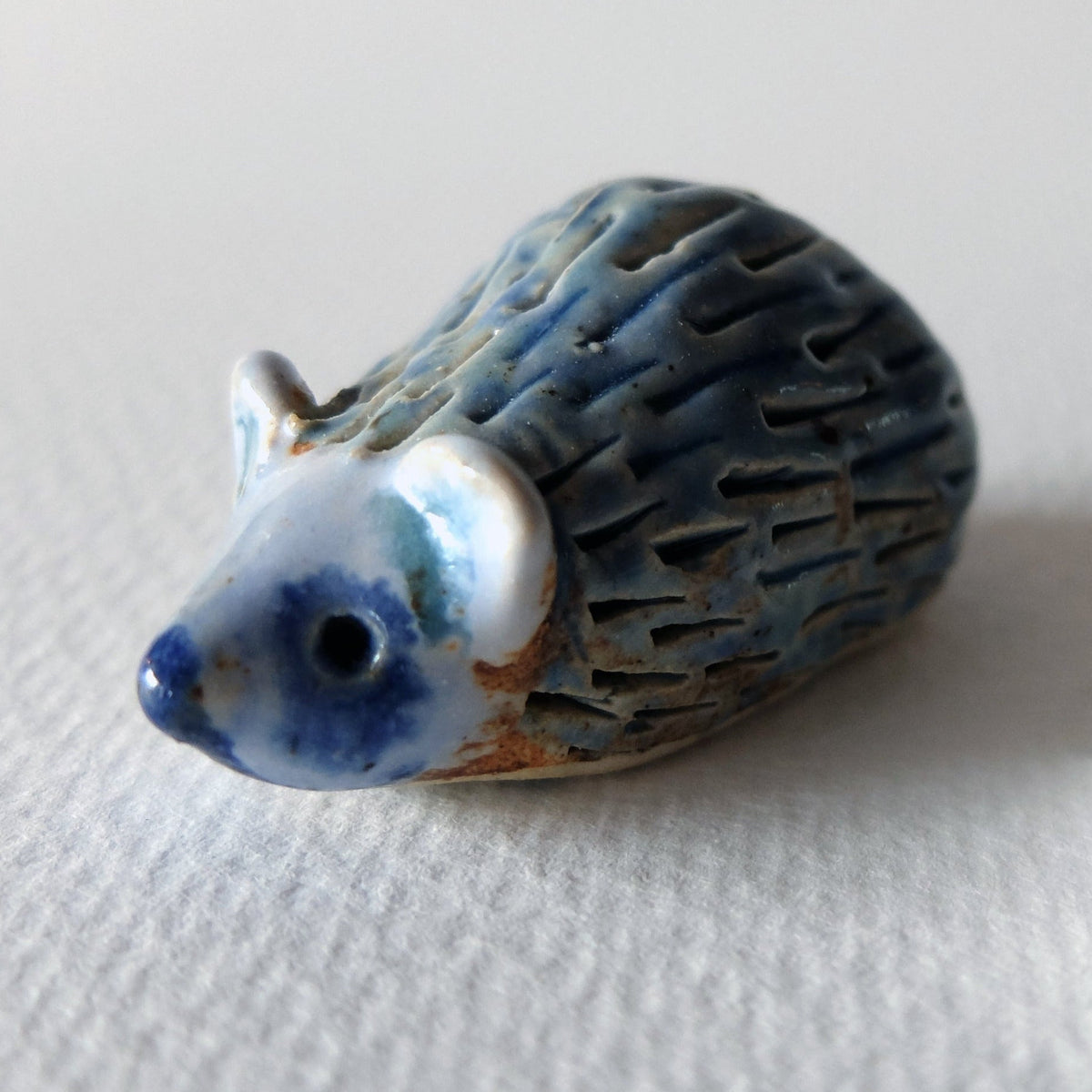 Handcrafted ceramic hedgehog figurine "The Timid Jade-Gazer (#1)" in stoneware by Nikolaj Jesper Cyon. Glazed with a shiny blue, white, and turquoise glaze on a terracotta stoneware body, with a textured pattern of indentations suggesting quills.